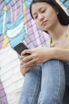 Young smiling woman sitting against a wall with graffiti and looking down and texting on her phone