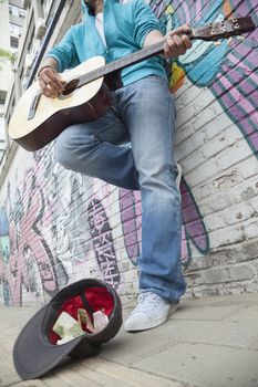 Young street musician playing guitar and busking for money in front of a wall with graffiti