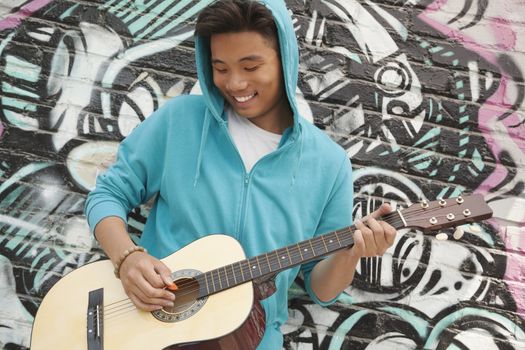 Young smiling street musician leaning on a wall with graffiti drawings and playing his guitar