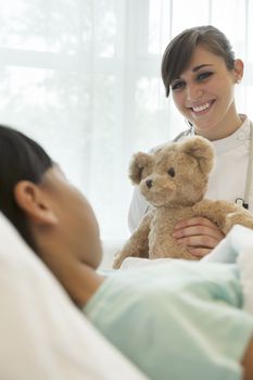 Smiling female doctor giving a teddy bear to a girl patient lying down on a hospital bed
