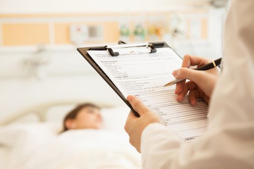 Close up of doctor writing on a medical chart with patient lying in a hospital bed in the background