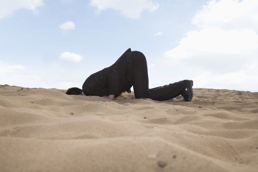 Young businessman kneeling with his head in a hole in the sand