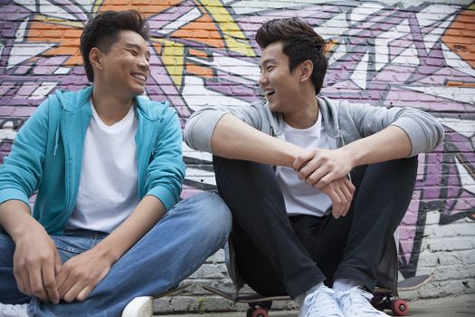 Two young men sitting on their skateboards and hanging out in front of a wall with graffiti