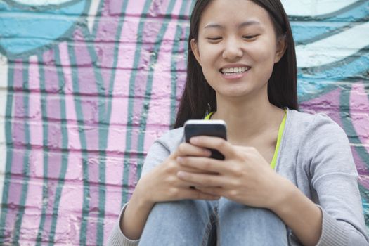 Young smiling woman sitting against a wall with graffiti and looking down and texting on her phone
