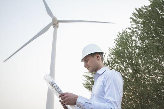 Young male engineer in a hardhat looking down at a blueprint in front of a wind turbine