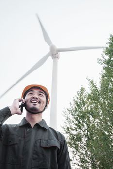 Young smiling male engineer in a hardhat on the phone beside a wind turbine