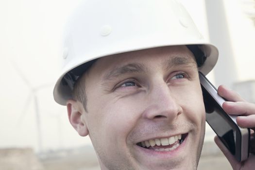 Young smiling male engineer in a hardhat on the phone beside a wind turbine, close-up