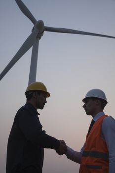 Two young male engineers standing beside a wind turbine at sunset and shaking hands