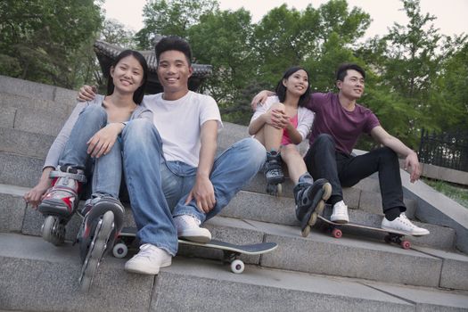 Two young couples sitting and resting on concrete steps outside with skateboards and roller blades
