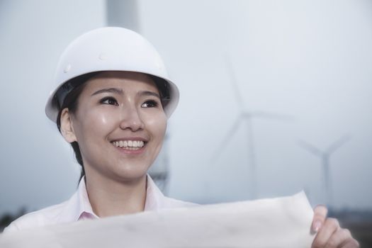 Young smiling female engineer holding a blueprint with wind turbines in the background