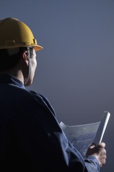 Young male engineer in a hardhat holding a blueprint at dusk, rear view