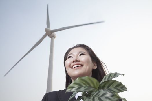 Portrait of smiling young businesswoman standing by a wind turbine and holding a plant