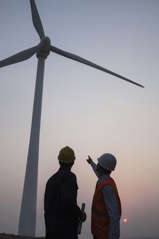 Two young male engineers standing beside a wind turbine at sunset, pointing up 