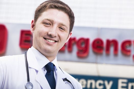 Portrait of young smiling doctor outside of the hospital, emergency room sign in the background