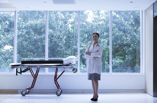Female Doctor with arms crossed standing next to a stretcher in the hospital, full length