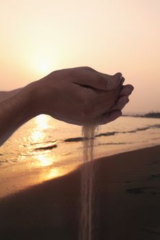 Hands holding and spilling sand with beach at sunset in the background