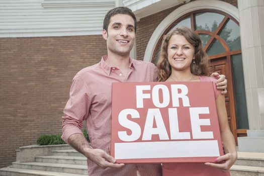 Smiling young couple holding a For Sale sign