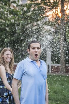 Couple playing with a garden hose and spraying each other outside in the garden, man has a shocked look 