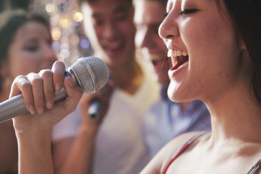 Close- up of young woman holding a microphone and singing at karaoke, friends singing in the background