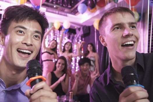 Two friends holding microphones and singing together at karaoke, friends in the background