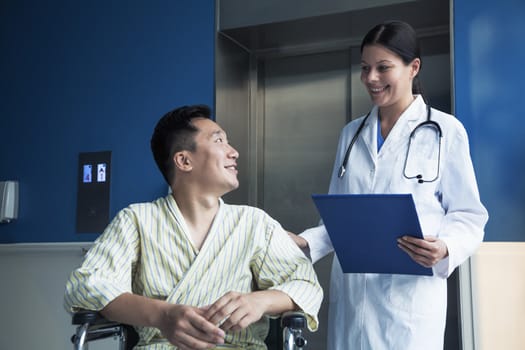 Young smiling male patient sitting in a wheelchair, looking up at the doctor standing beside him