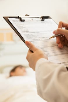 Close up of doctor writing on a medical chart with patient lying in a hospital bed in the background