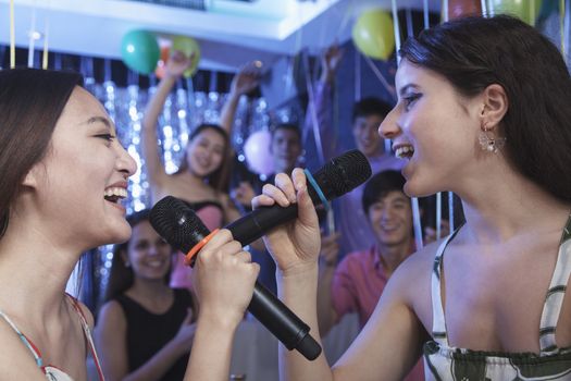 Two friends holding microphones and singing together at karaoke, face to face, friends in the background