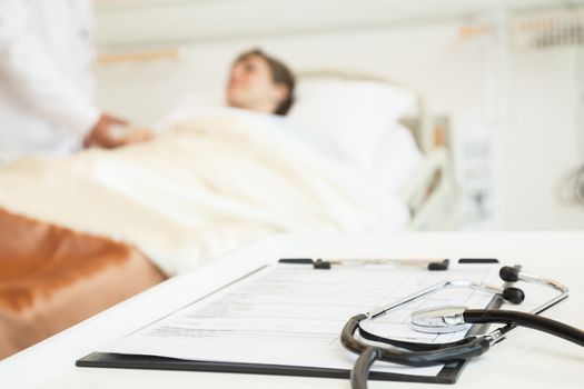 Close-up of stethoscope and medical chart with a patient lying in a hospital bed in the background