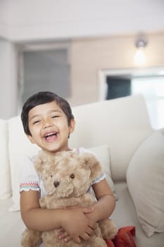 Happy, smiling girl holding a teddy bear and sitting on the couch, looking at camera