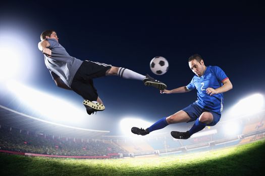 Two soccer players in mid air kicking the soccer ball, stadium lights at night in background