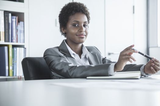 Portrait of young smiling businesswoman sitting at a table and holding a pen, looking at camera