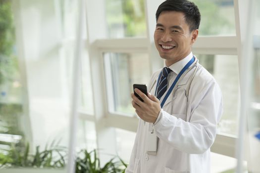 Smiling doctor using his phone in the hospital lobby, looking at camera, glass doors