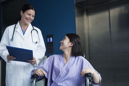 Young smiling female patient sitting in a wheelchair, looking up at the doctor standing beside her