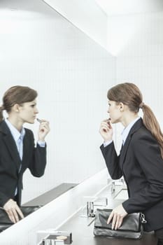 Young businesswoman with long hair looking into the mirror and putting on lipstick in the bathroom