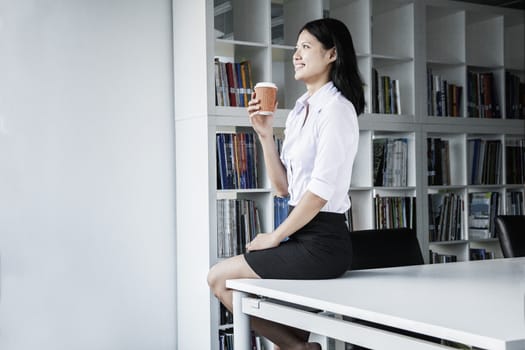 Smiling young businesswoman holding a coffee and sitting on a table, looking away