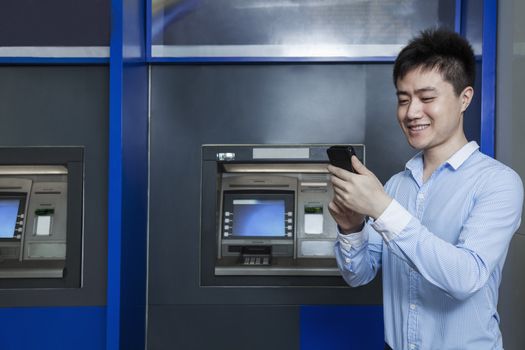 Smiling young businessman standing in front of an ATM and looking at his phone