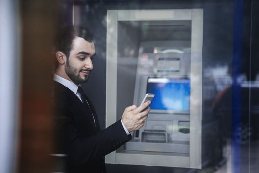 Smiling young businessman standing in front of an ATM and looking at his phone