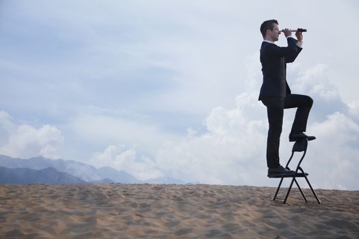 Businessman standing on a chair and looking through a telescope in the middle of the desert 