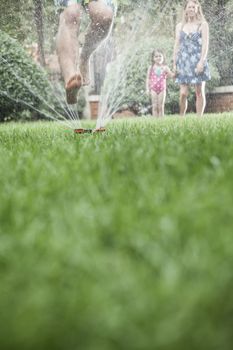 Surface level shot of father jumping through a sprinkler in the grass, mother and daughter watch in the background