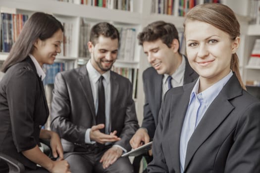 Business woman smiling and looking at the camera with her colleagues talking and looking down at a digital tablet in the background