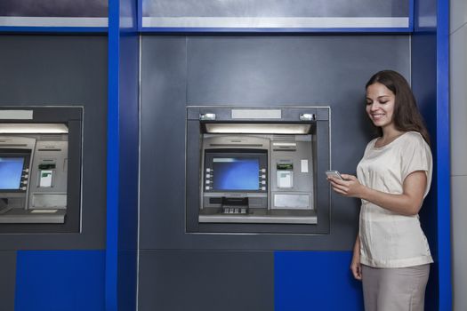 Smiling young woman standing in front of an ATM and looking at her phone