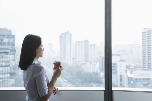 Smiling young businesswoman holding a coffee cup and looking out the window a the cityscape in Beijing, China