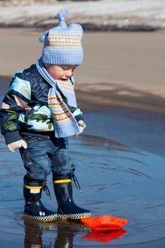 Little boy plays with paper ships in a spring puddle