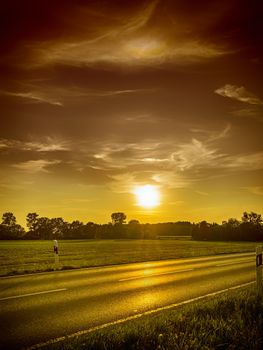 Picture of a road at sunset with backlight, meadows and trees