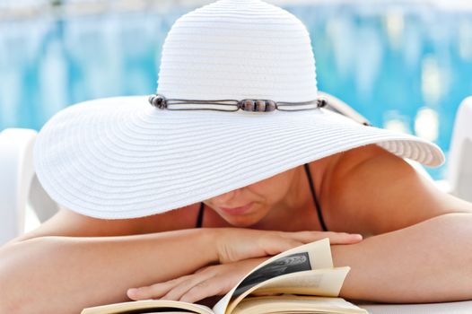 girl in a white hat reading a book in a lounge chair by the pool