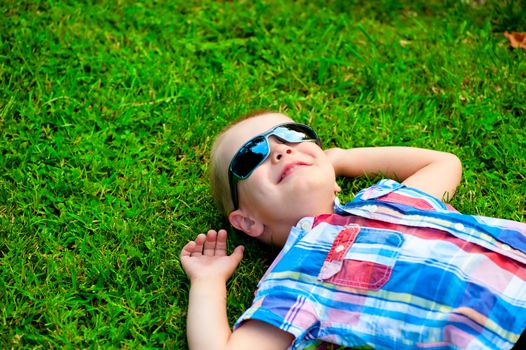 happy little boy lying down resting on the green grass in sunglasses