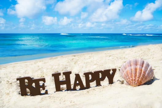 Sign "Be Happy" and seashell on the sandy beach by the ocean in sunny day