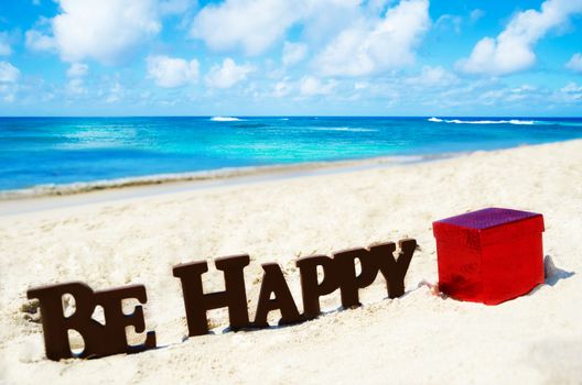 Sign "Be Happy" and gift box on the sandy beach by the ocean in sunny day