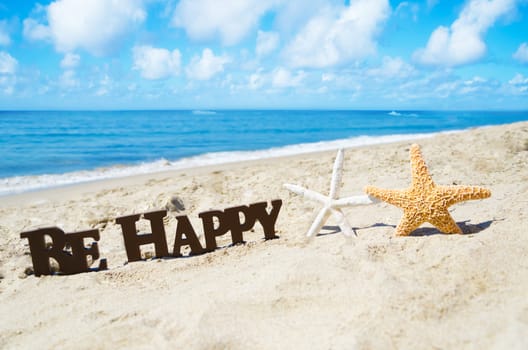 Sign "Be Happy" and two starfishes on the sandy beach by the ocean in sunny day