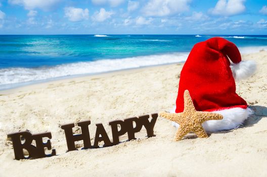 Sign "Be Happy" and starfish with christmas hat on the sandy beach by the ocean in sunny day